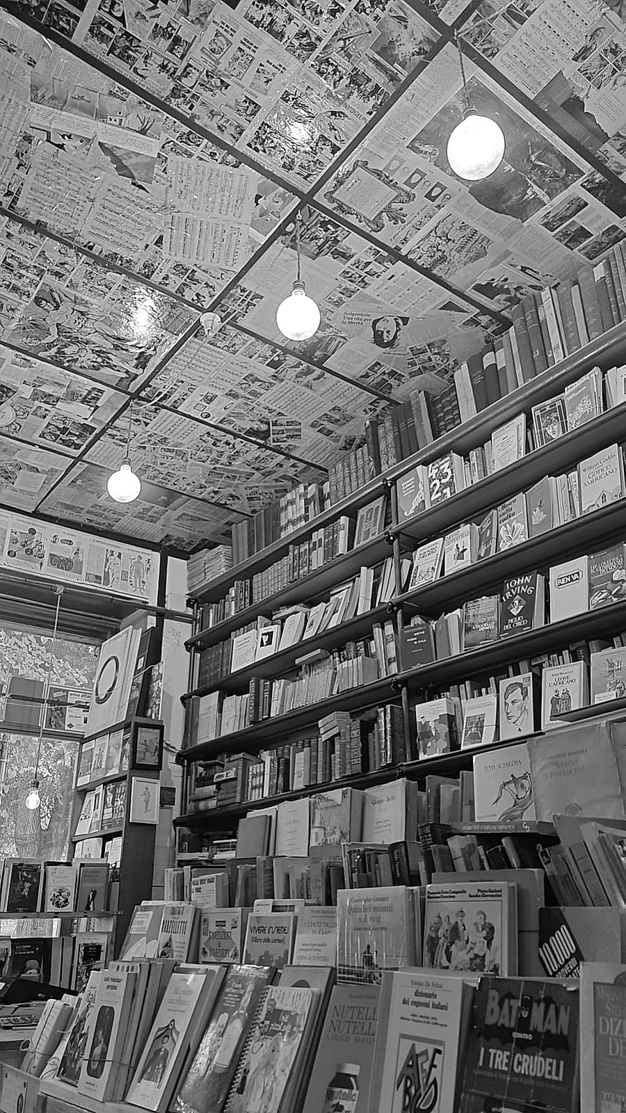 L'interno della libreria: scaffali carichi di libri fino al soffitto tappezzato di pagine a fumetti, con le lampadine appese.